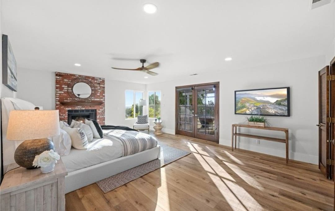Spacious modern bedroom with a brick fireplace and large windows, featuring stylish decor.