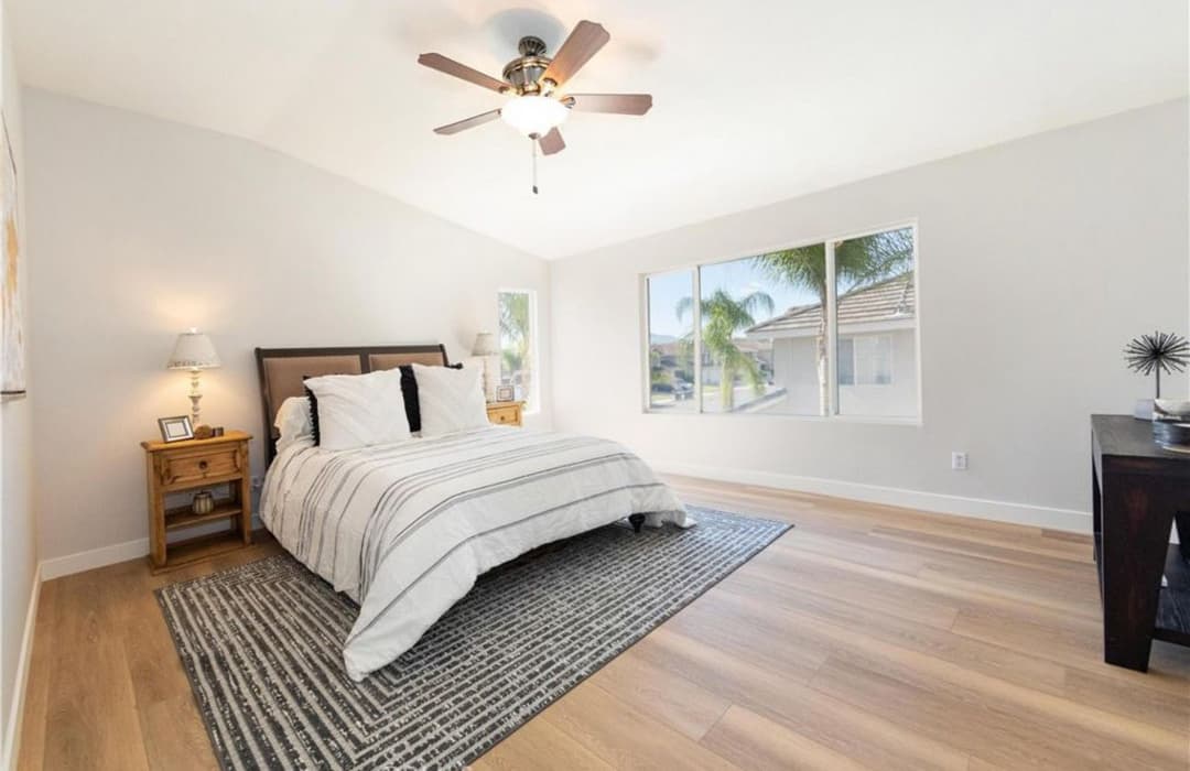 Modern bedroom with a queen bed, ceiling fan, large windows, and hardwood flooring.