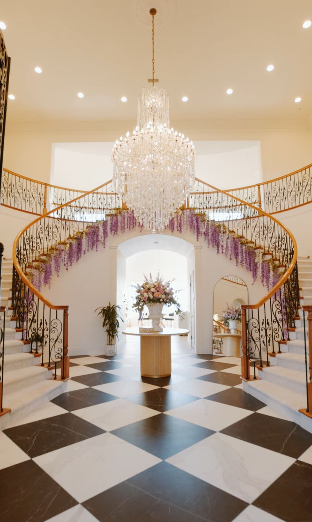 Elegant foyer with a grand chandelier and spiral staircase adorned with purple wisteria.