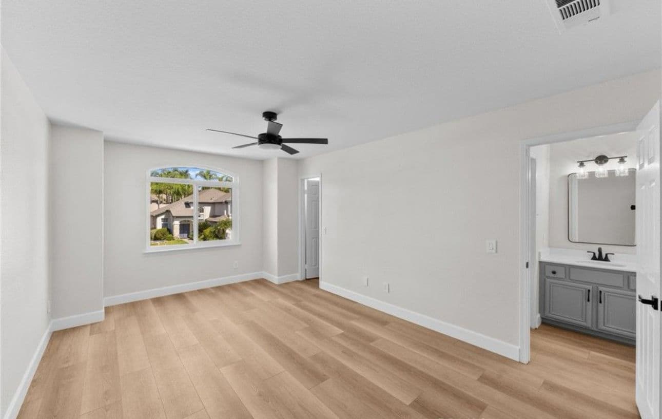 Spacious bedroom with white walls, large window, light wood flooring, and modern ceiling fan.