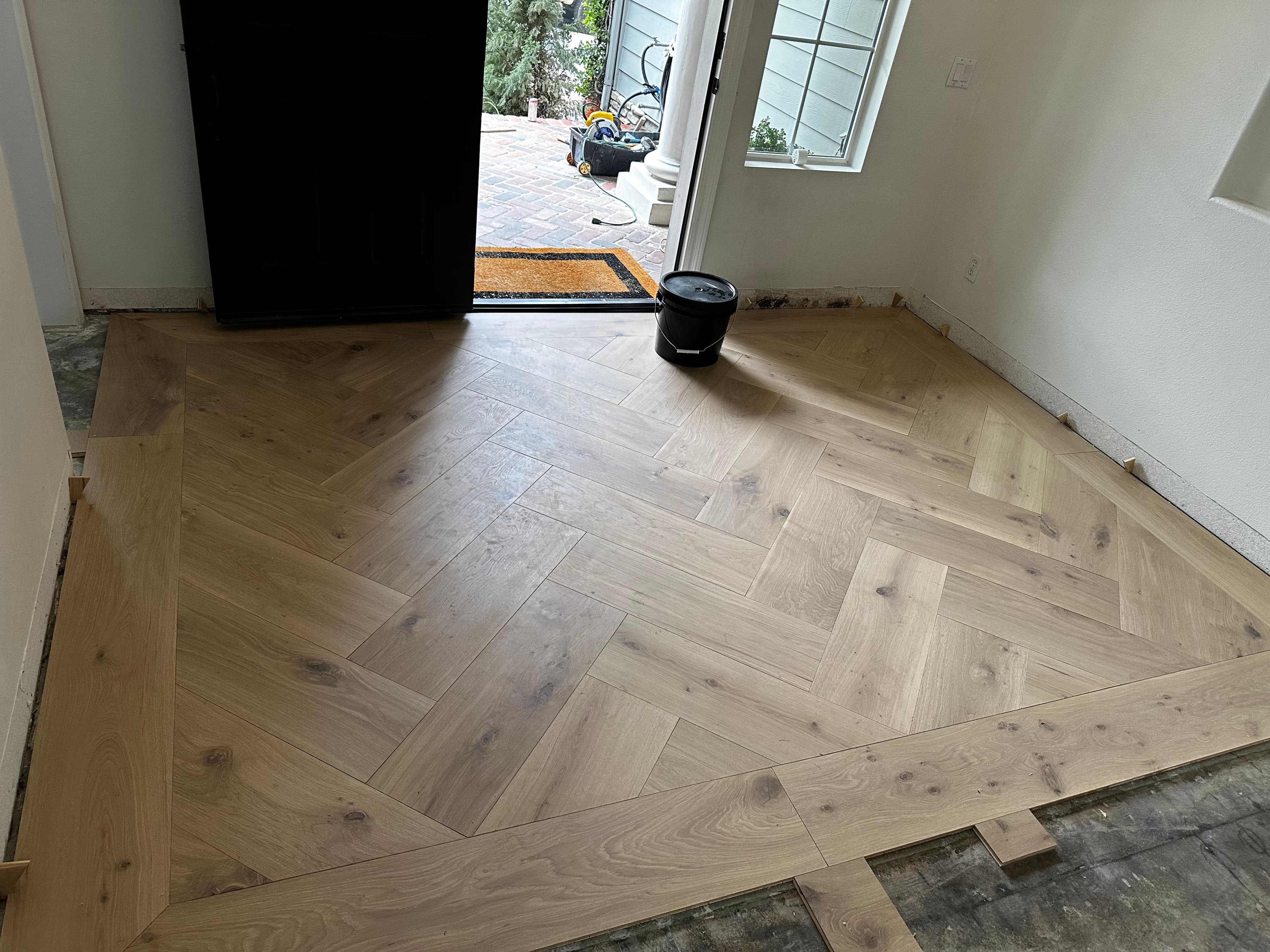 Herringbone wood flooring installation with an open door and bucket in a home entryway.